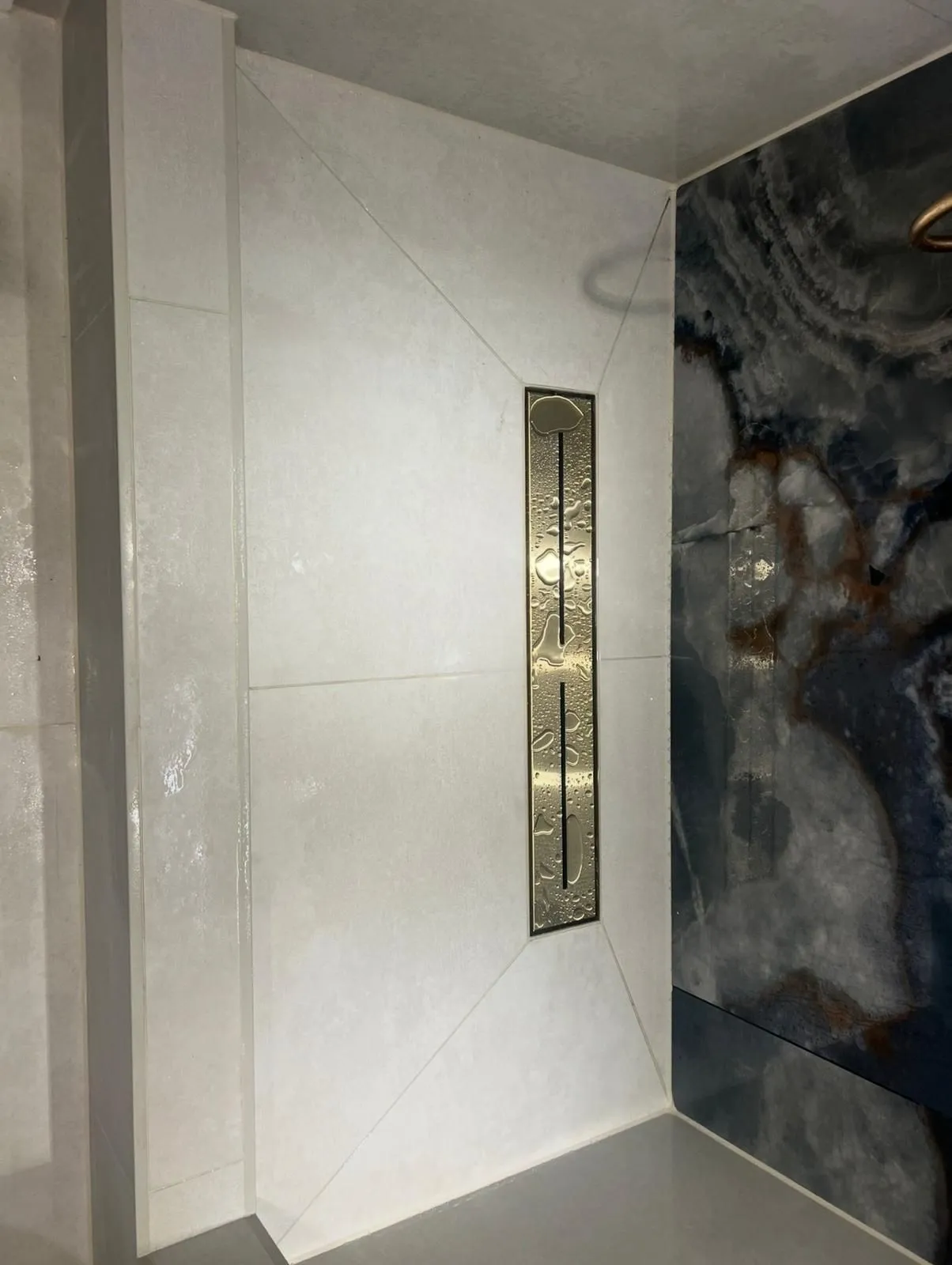 Onyx-effect slab beside a light porcelain shower wall with a brass linear drain